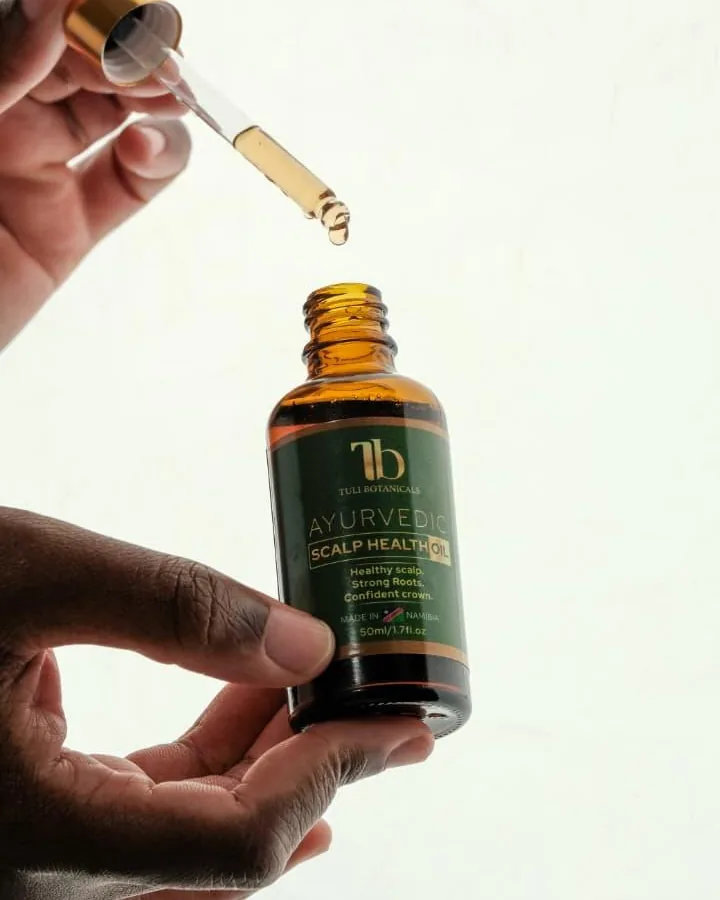 Golden oil dripping from a glass dropper into an open bottle of Tuli Botanicals Scalp Health Oil.