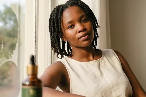 Seated by a window with natural hair, bottle in hand.